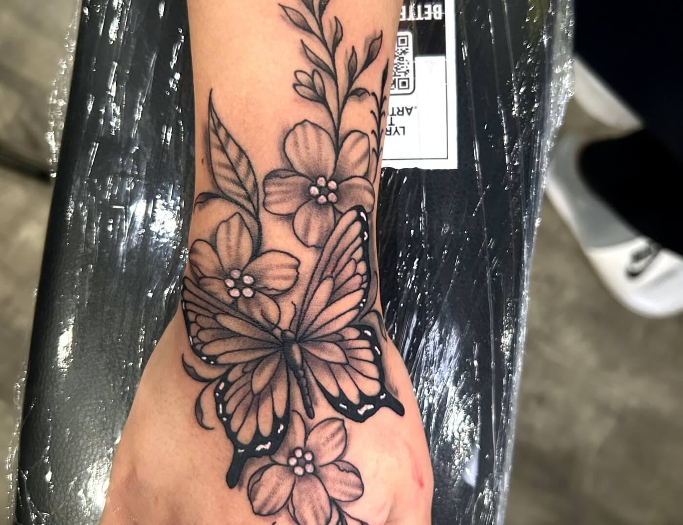 Butterfly among flowers tattoo in Black & Grey By Lyric TheArtist at Iron Palm Tattoos In Atlanta, Georgia. Butterflies and flowers complement each other aesthetically in the real world and in art. The delicate, colorful nature of butterflies can be enhanced by the addition of vibrant, detailed flowers. Additionally, butterflies and flowers are often associated with spring and renewal. Call 404-973-7828 or stop by for a free consultation with Lyric or another Iron Palm Tattoo body artist. Walk-ins are welcome.