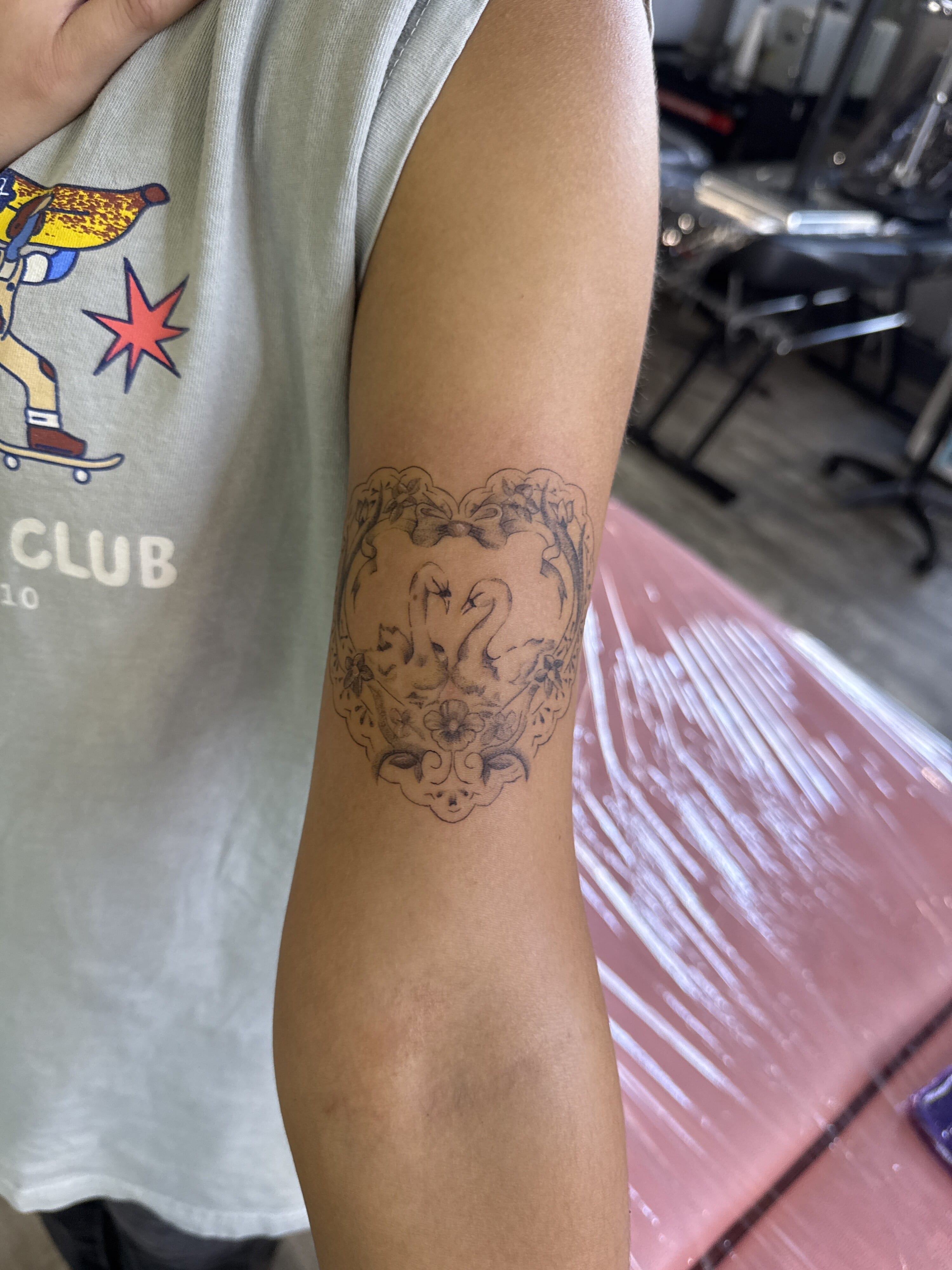 A delicate Fine Line tattoo features two swans forming a heart-shaped composition, surrounded by flowers and ornamental details. The soft Black & Gray linework gives the design an elegant, romantic finish. Tattoo by Dash. Made at Iron Palm Tattoos & Body Piercings in downtown Atlanta, GA. Call or text 404-973-7828. Tattoo consultations are free and walk ins are always welcome.